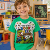 A young boy smiling in a colorful children's classroom, wearing a bright green graphic t-shirt that reads 'THIS IS MY BOOK WEEK COSTUME!' with a dinosaur illustration.