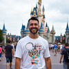 Man wearing a Disney 2026 t-shirt in front of a castle at a theme park.