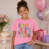 A smiling girl wearing a pink graphic t-shirt that reads 'HAPPY DOUBLE BIRTHDAY ERA' in colorful bubble letters, standing in a birthday-themed room with balloons, a cake, and wrapped gifts.
