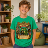 A smiling boy wearing a bright green graphic T-shirt in a cozy indoor room with bookshelves, plants, and a beanbag chair. The shirt features a detailed fantasy illustration with a dragon, castle, scrolls, and the text 'A Book Week Quest: Hayden Dragon Valley'.