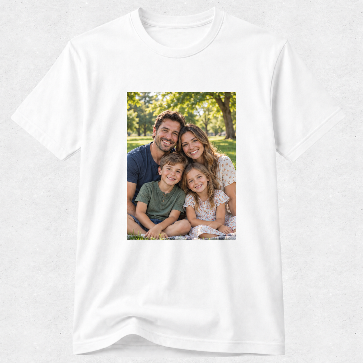 A white short-sleeve crewneck t-shirt with a rectangular custom photo print on the chest showing a family of four in a park, displayed on a light gray textured background.