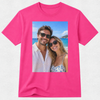 A bright pink short-sleeve crewneck t-shirt with a rectangular graphic print on the front showing a smiling couple posing on a sunny beach.