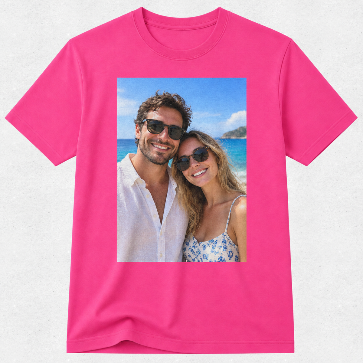 A bright pink short-sleeve crewneck t-shirt with a rectangular graphic print on the front showing a smiling couple posing on a sunny beach.