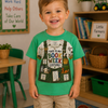 A young boy smiling in a classroom setting, wearing a bright green graphic t-shirt that reads 'THIS IS MY BOOK WEEK COSTUME!' with a book-and-costume illustration, paired with khaki cargo shorts.