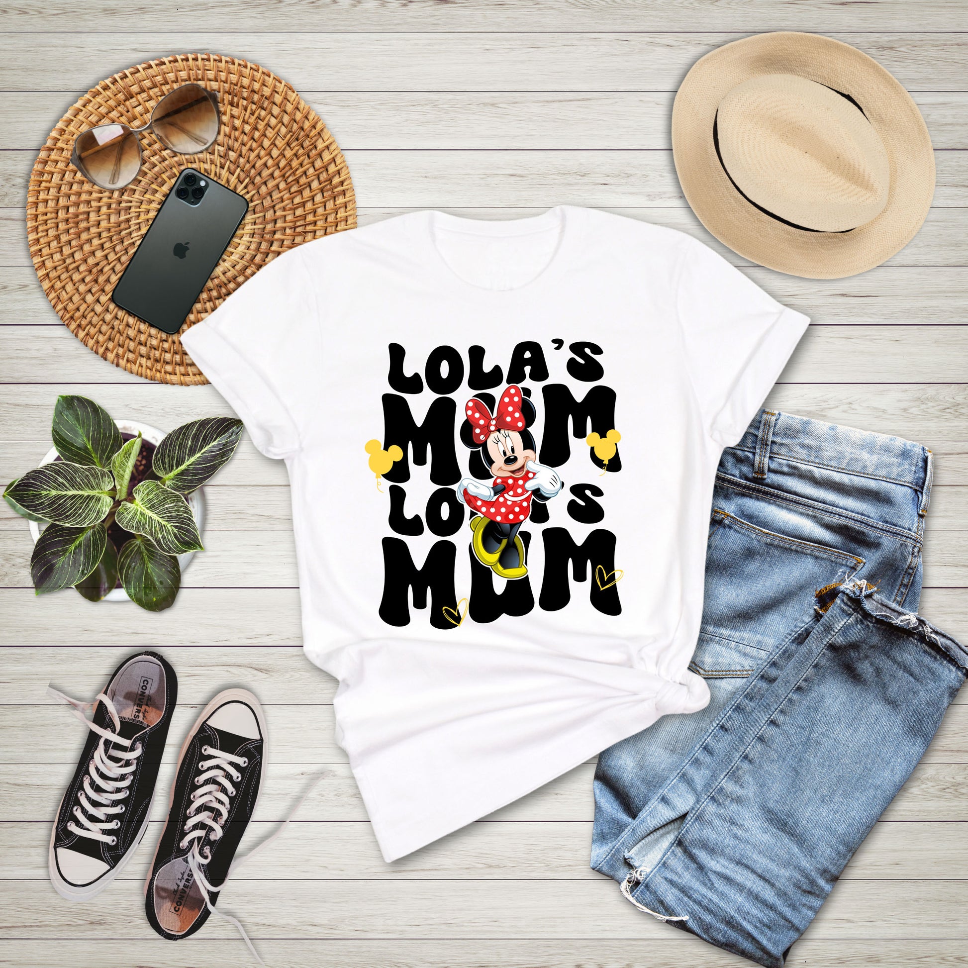 White t-shirt with 'LOLA'S MOM LOLO IS MOM' text and Minnie Mouse graphic, surrounded by jeans, sneakers, a hat, and phone on a wooden surface.