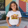 A young girl smiling and wearing a white graphic t-shirt that reads 'It's OKAY TO NOT BE OKAY' with a rainbow and sun design, set against a colorful outdoor wall.