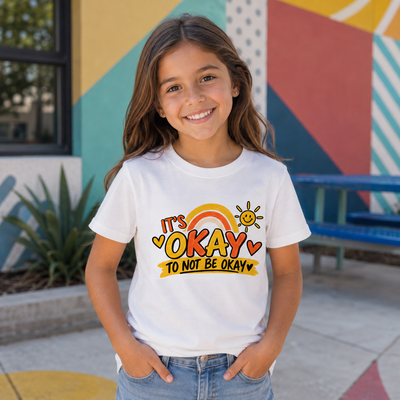 A young girl smiling and wearing a white graphic t-shirt that reads 'It's OKAY TO NOT BE OKAY' with a rainbow and sun design, set against a colorful outdoor wall.