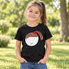 A young girl with a ponytail stands outdoors on a grassy lawn wearing a black t-shirt with a cartoon graphic of a girl with brown hair and a red hair tie, paired with blue jeans.