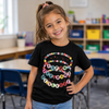 A young girl with a pink hair bow smiling in a classroom, wearing a black t-shirt with a colorful 'Happy 100 Days of School' banner graphic.