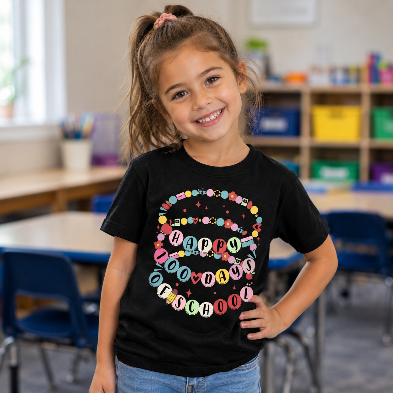 A young girl with a pink hair bow smiling in a classroom, wearing a black t-shirt with a colorful 'Happy 100 Days of School' banner graphic.