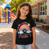 A young girl smiling outdoors in front of a school playground and building, wearing a black graphic T-shirt that reads 'HAPPY 100TH DAY OF SCHOOL' with a colorful rainbow, clouds, hearts, and stars.