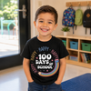 A young boy smiling in a school classroom, wearing a black graphic t-shirt that reads 'HAPPY 100 DAYS OF SCHOOL' with a rainbow and pencil design.
