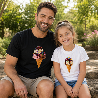 A man and a young girl sitting together outdoors on a stone path, wearing matching custom ice cream cone graphic t-shirts. The man wears a black t-shirt and the girl wears a white t-shirt, both featuring a detailed graphic of a vanilla ice cream scoop in a waffle cone with chocolate drizzle and sprinkles.
