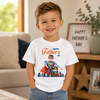 A young boy smiling in a bright living room, wearing a white short-sleeve graphic T-shirt that says 'Happy Father's Day' with two superhero characters on the front, paired with blue jeans.