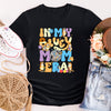 Black t-shirt with colorful graphic design, surrounded by white sneakers, a straw hat, and denim shorts on a light background.