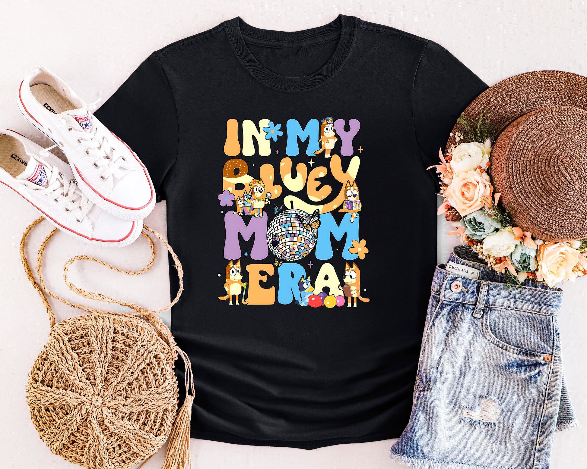 Black t-shirt with colorful graphic design, surrounded by white sneakers, a straw hat, and denim shorts on a light background.