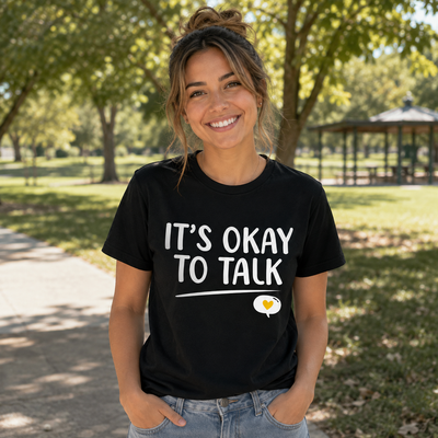 A woman smiling outdoors in a black graphic t-shirt that reads 'IT'S OKAY TO TALK' with a small heart-shaped bubble graphic below, paired with light blue jeans in a park setting.