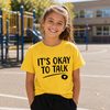 A young girl smiling in a yellow graphic t-shirt that reads 'IT'S OKAY TO TALK' with a small heart icon, standing on an outdoor playground.