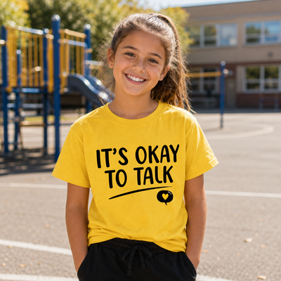 A young girl smiling in a yellow graphic t-shirt that reads 'IT'S OKAY TO TALK' with a small heart icon, standing on an outdoor playground.