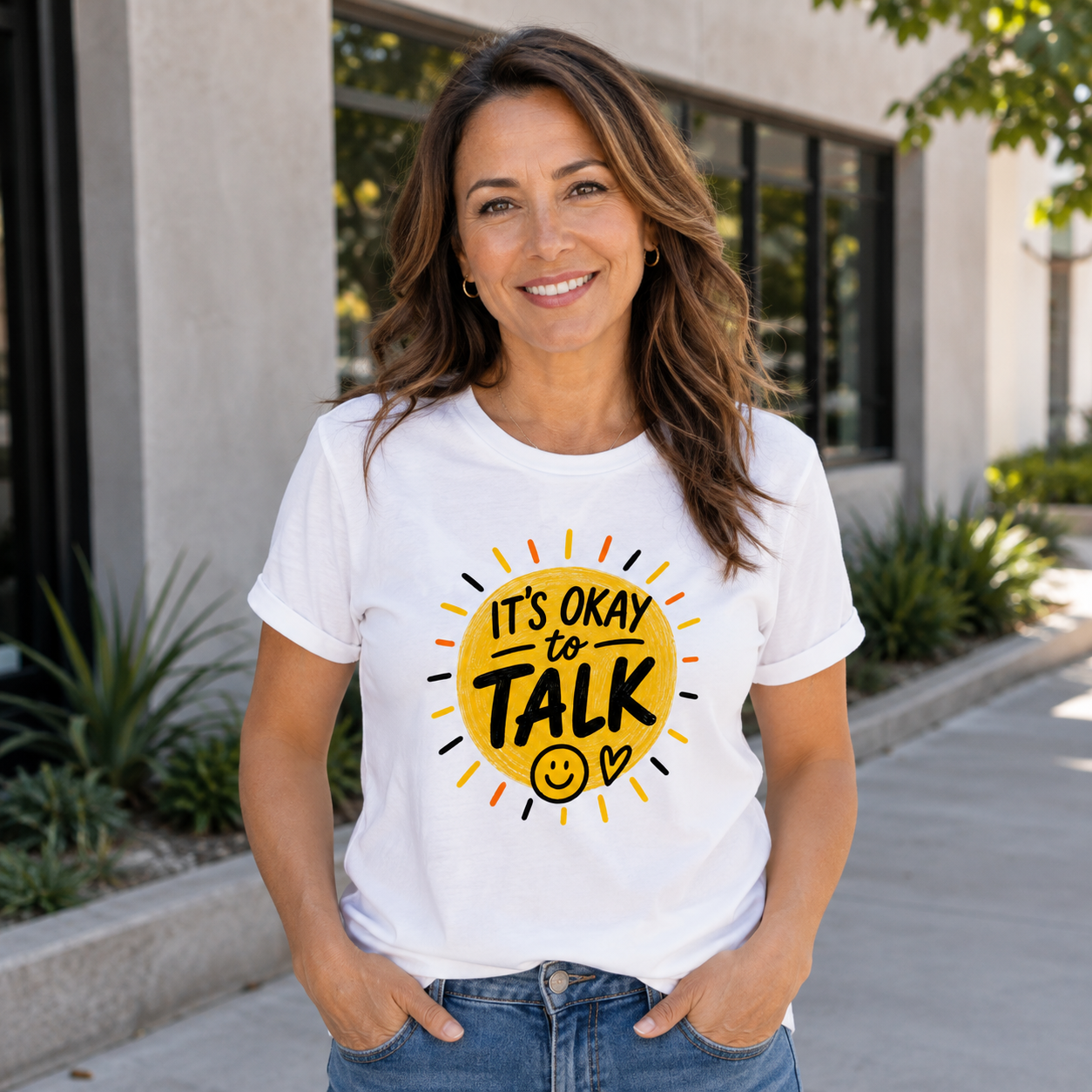 A woman wearing a white graphic t-shirt with a yellow sun design that reads 'IT'S OKAY TO TALK' with a smiley face, standing outdoors in front of a modern building.