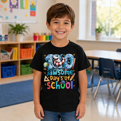A smiling young boy stands in a classroom wearing a black graphic t-shirt that reads '100 Jawsome Days of School' with colorful lettering and school-themed illustrations.