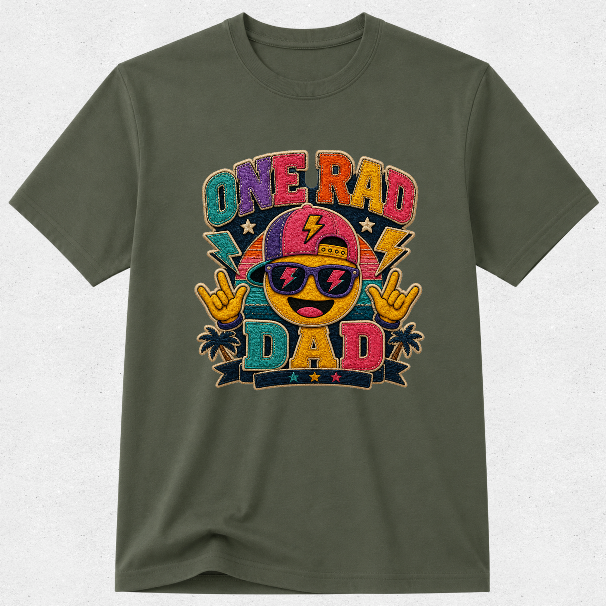 An olive green short-sleeve t-shirt with a colorful graphic on the front that reads 'ONE RAD DAD' with a smiling emoji face wearing sunglasses and a pink cap, set against a light gray textured background.