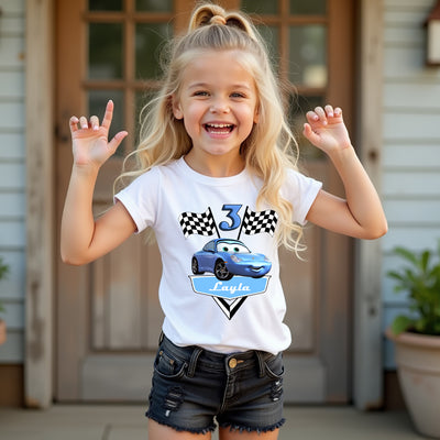 Young girl wearing a white t-shirt with a car design and number 3, standing outside a house.