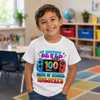 A smiling young boy wearing a white graphic t-shirt with a colorful 'LEVEL 100 DAYS OF SCHOOL UNLOCKED' design, standing in a brightly lit classroom.