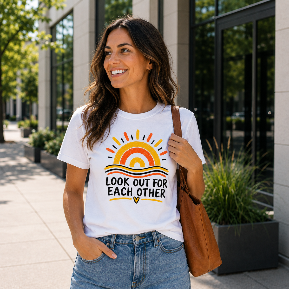 A woman wearing a white graphic t-shirt with a colorful sun and the text 'LOOK OUT FOR EACH OTHER' printed on the front, paired with blue jeans and a brown shoulder bag, standing outdoors in front of a modern building.
