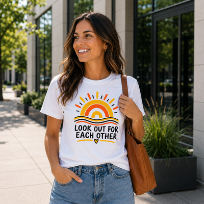A woman wearing a white graphic t-shirt with a colorful sun and the text 'LOOK OUT FOR EACH OTHER' printed on the front, paired with blue jeans and a brown shoulder bag, standing outdoors in front of a modern building.