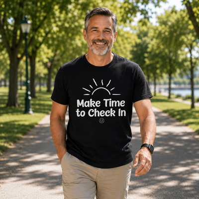 A smiling man walking on a tree-lined path wearing a black t-shirt with a white graphic that reads 'Make Time to Check In' and khaki pants.