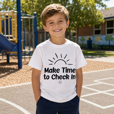 A smiling young boy stands on a playground wearing a white graphic t-shirt with a black sun illustration and the text 'Make Time to Check In'.