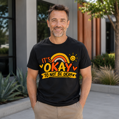 A smiling man wearing a black graphic T-shirt that reads 'IT'S OKAY TO NOT BE OKAY' with a rainbow, sun, and heart details, standing outdoors in front of a modern building.