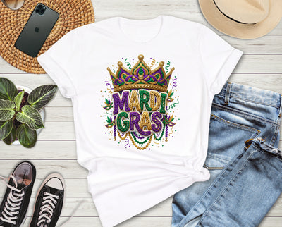 White t-shirt with Mardi Gras design on a wooden surface with jeans, sneakers, and a hat.