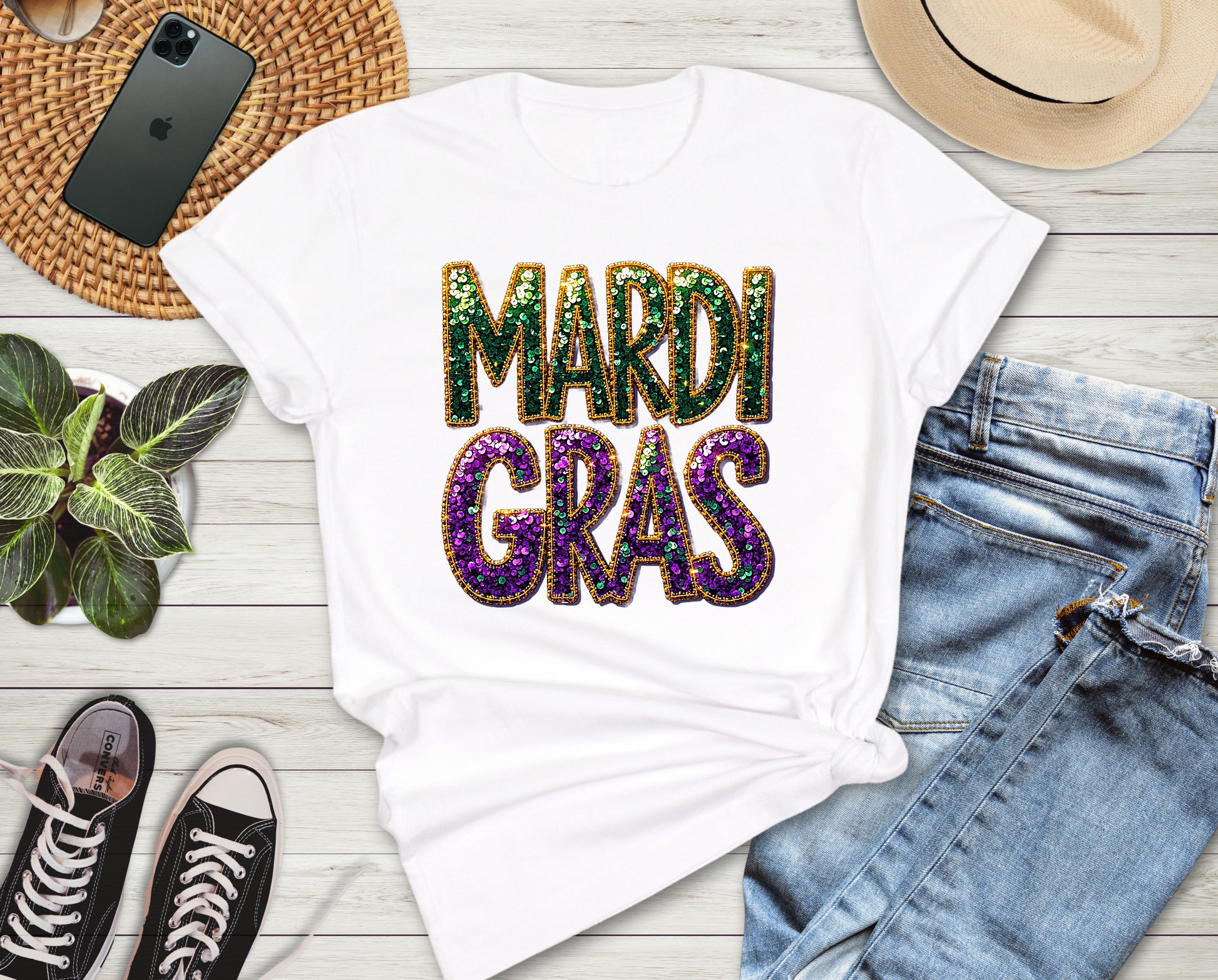 White t-shirt with 'Mardi Gras' text on a wooden surface with jeans, sneakers, and a hat.