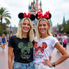 Two women wearing Disney-themed shirts and ears in front of a castle.
