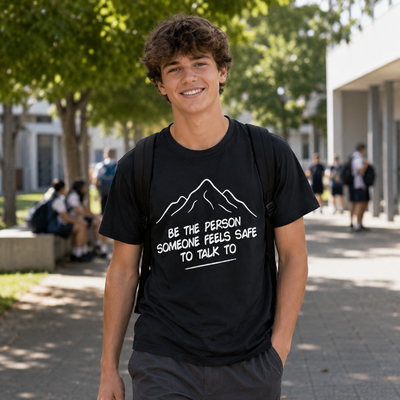 A smiling young man standing outdoors on a campus walkway wearing a black graphic t-shirt with a white mountain outline and the text 'BE THE PERSON SOMEONE FEELS SAFE TO TALK TO'.
