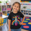 A young girl with braided hair smiling in a colorful school classroom, wearing a black t-shirt with a vibrant graphic of a pencil, hearts, and a rainbow that reads 'HAPPY 100 DAYS OF SCHOOL'.