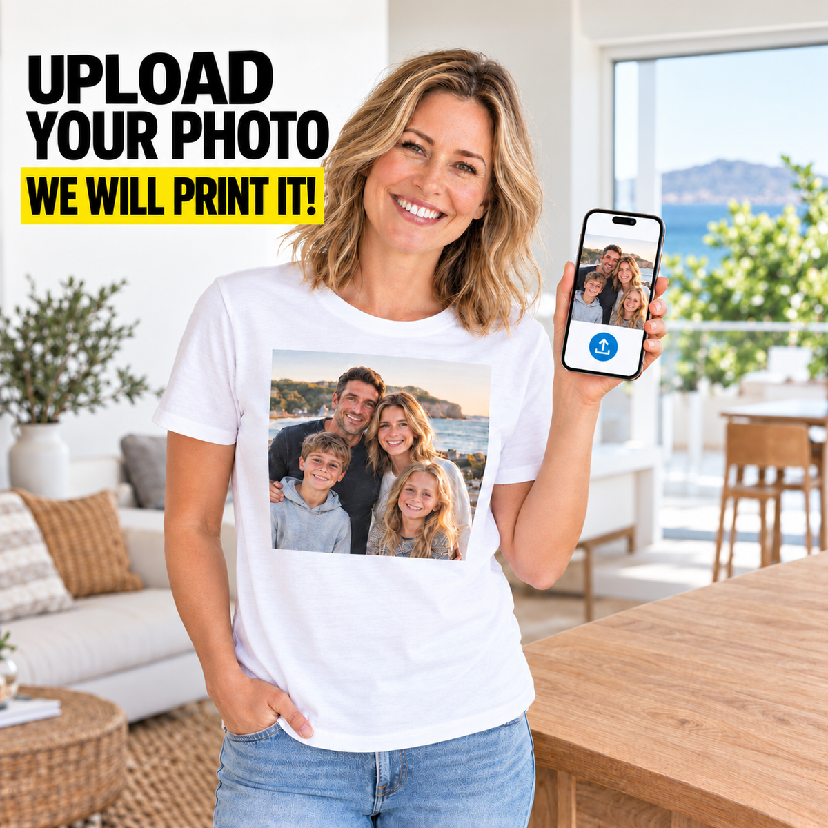 A smiling woman stands in a bright living room wearing a white T-shirt with a printed family photo on the front, while holding up a smartphone that shows the photo being used for the print. Large text in the upper left reads 'UPLOAD YOUR PHOTO WE WILL PRINT IT!'.