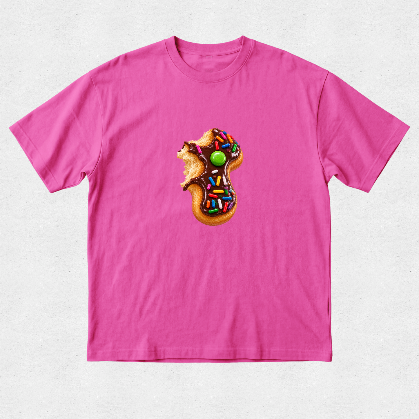 A bright pink short-sleeve crewneck t-shirt featuring a central graphic of a donut-shaped cookie decorated with chocolate frosting, colorful sprinkles, and a green candy, with a small brown bear sitting on top.