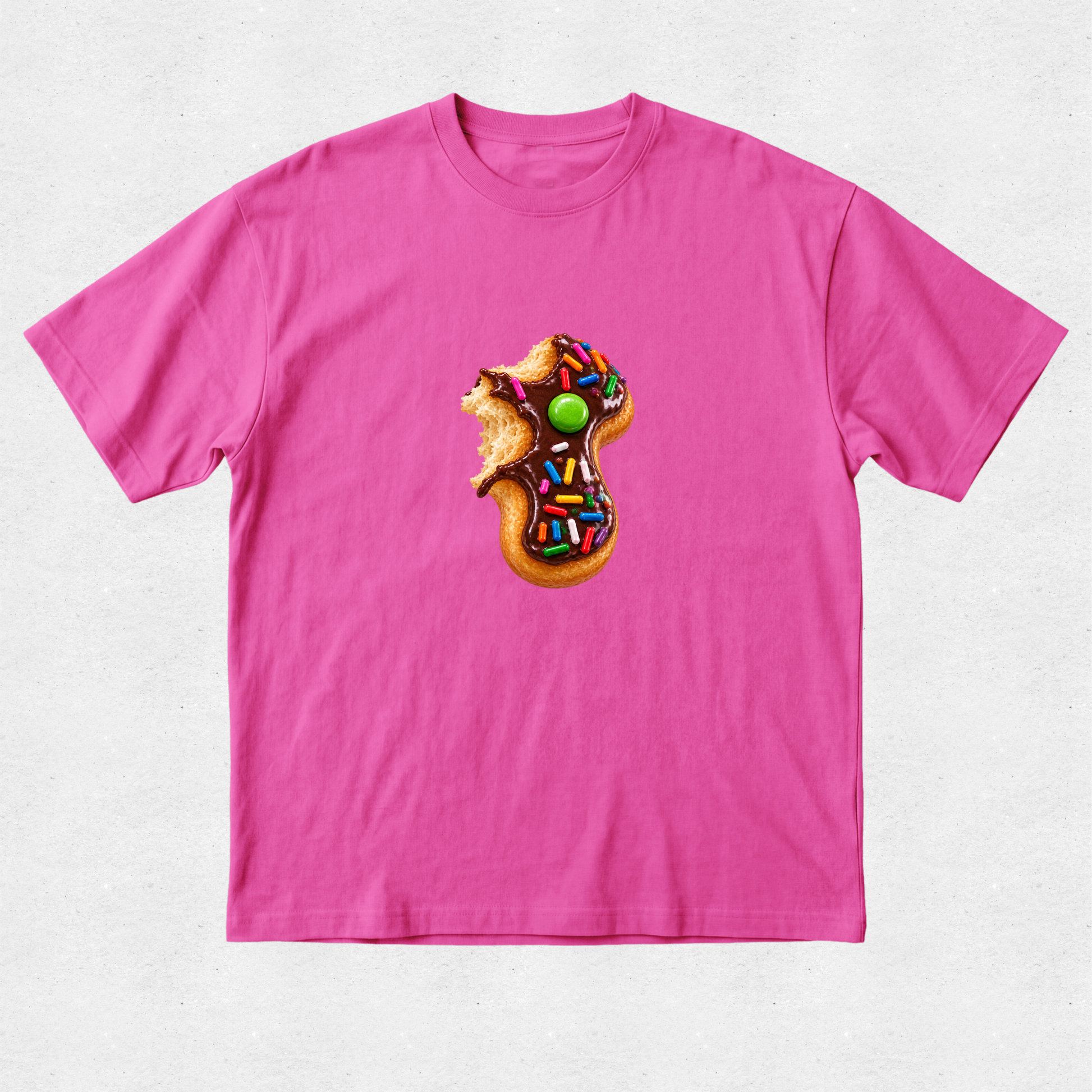 A bright pink short-sleeve crewneck t-shirt featuring a central graphic of a donut-shaped cookie decorated with chocolate frosting, colorful sprinkles, and a green candy, with a small brown bear sitting on top.