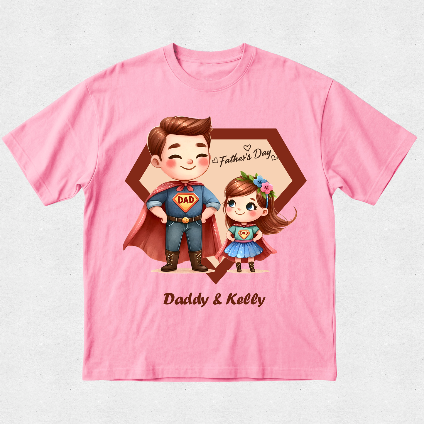 A pink short-sleeve crewneck t-shirt featuring a custom cartoon graphic of a father and daughter in superhero costumes, with 'Father's Day' and 'Daddy & Kelly' printed on the front.