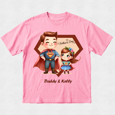 A pink short-sleeve crewneck t-shirt featuring a custom cartoon graphic of a father and daughter in superhero costumes, with 'Father's Day' and 'Daddy & Kelly' printed on the front.