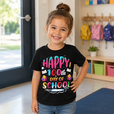 A young girl with her hair in a bun stands indoors wearing a black graphic T-shirt that reads 'HAPPY 100th DAY OF SCHOOL' in colorful lettering, paired with blue jeans. The background shows a classroom-like interior with backpacks hanging on a rack and storage bins.