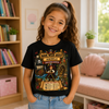 A young girl with curly brown hair in a ponytail smiles while wearing a black graphic t-shirt with a pirate-themed 'Book Week Quest' design, paired with light blue jeans in a cozy, sunlit room with bookshelves and a window.