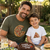 A smiling man and young boy sitting together outdoors at a table, each wearing a pizza-themed graphic T-shirt. The man wears an olive green shirt with a large sliced pizza graphic, and the boy wears a white shirt with a smaller pizza slice graphic.
