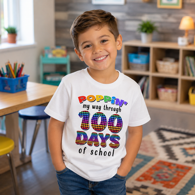 A young boy smiling in a classroom setting, wearing a white graphic t-shirt that reads 'POPPIN' my way through 100 DAYS of school' in colorful, patterned lettering.