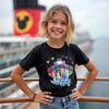 Young girl wearing a black t-shirt with a colorful graphic design, standing on a pier with a disney cruise ship in the background.