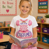 A young girl with blonde hair in a ponytail stands in a classroom holding a book, wearing a white graphic T-shirt that says 'THIS IS MY BOOK WEEK COSTUME!' with a pink princess dress illustration, and a pink tutu skirt.