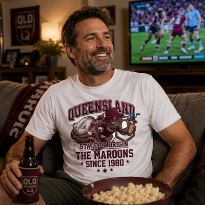 Man wearing a Queensland State of Origin t-shirt, holding a beer and popcorn, watching TV in a living room.
